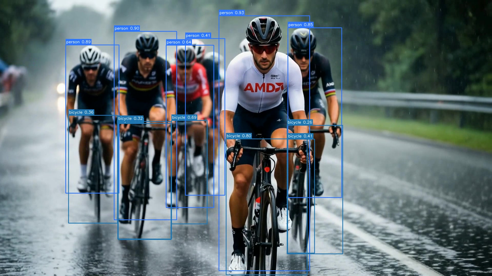 Frame at the 2-second mark of the sample clip showing YOLO26 bounding boxes drawn over a peloton of AMD-jersey cyclists on a rain-soaked road; person and bicycle classes detected with confidences from 0.26 to 0.93